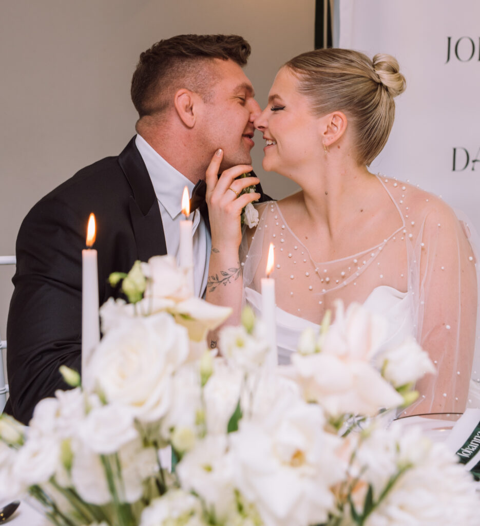 „Brautpaar küsst sich während des Hochzeitsdinners mit eleganter weißer Floristik bei einer Hochzeit in Ingolstadt.“