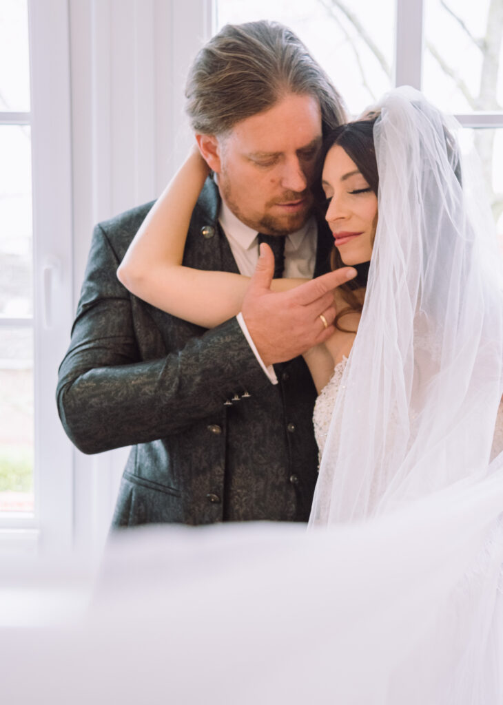 „Intimes Brautpaarportrait mit Schleier im Fensterlicht bei einer Hochzeit in Ingolstadt.“
