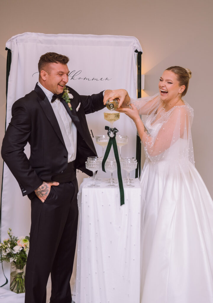 „Brautpaar gießt Champagner in einen Champagne Tower während der Hochzeitsfeier in Ingolstadt; Hochzeitsszene voller Freude.“